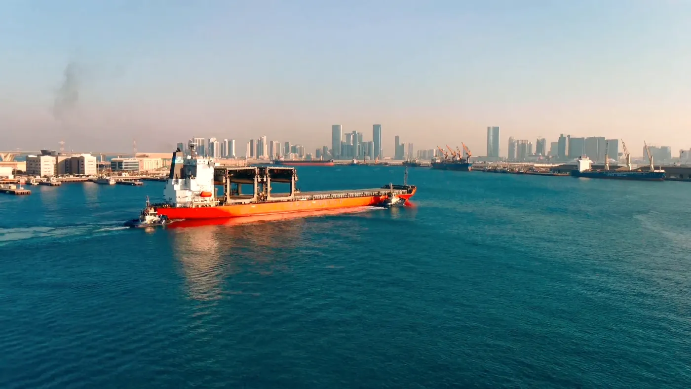 A cargo ship under tow in a harbour, with a city skyline behind it